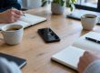 Smartphone en enregistrement posé sur une table de réunion en bois, entre carnets ouverts et tasses de café