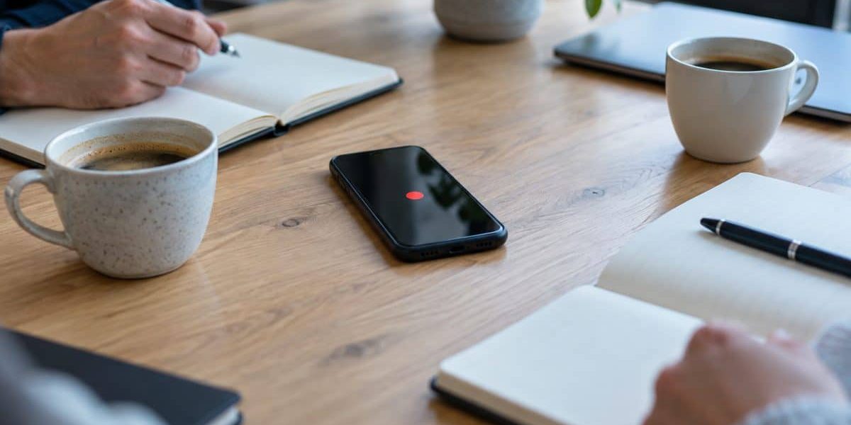 Smartphone en enregistrement posé sur une table de réunion en bois, entre carnets ouverts et tasses de café